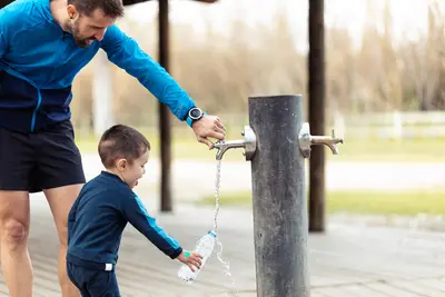 Vater öffnet Wasserhahn des Trinkbrunnes, während Kindergartenkind die Wasserflasche auffüllt.