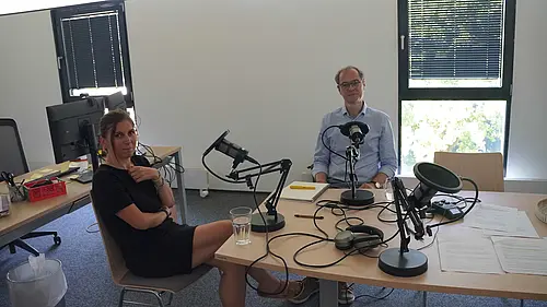 ein Mann und eine Frau vor Podcast-Mikrofonen lächeln in die Kamera
