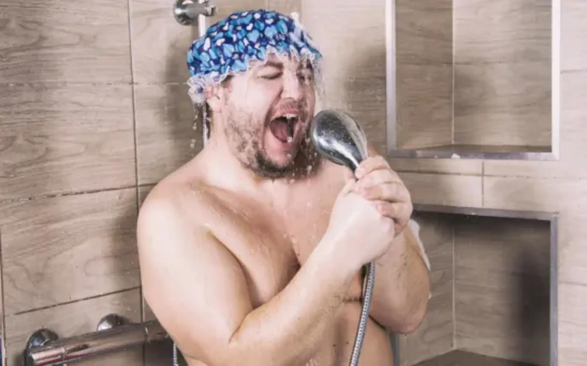 Ein Mann mit Duschhaube steht in der Dusche und singt emotional in den Duschkopf.