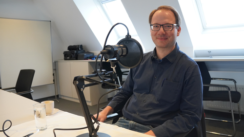 ein Mann sitzt vor einem Mikro. Er lächelt in die Kamera.