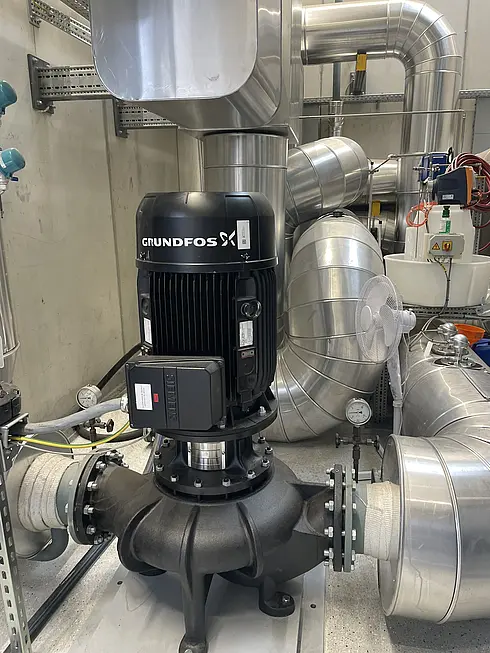 Industriepumpensystem mit einem Grundfos-Motor, der auf einem großen schwarzen Pumpengehäuse montiert ist. Das System ist mit mehreren Metallrohren und -leitungen verbunden. Rundherum sind Messgeräte und weitere technische Komponenten sichtbar, was auf eine größere mechanische oder HLK-Anlage hinweist. Die Umgebung wirkt wie ein Industrie- oder Technikraum für Flüssigkeits- oder Luftsysteme.