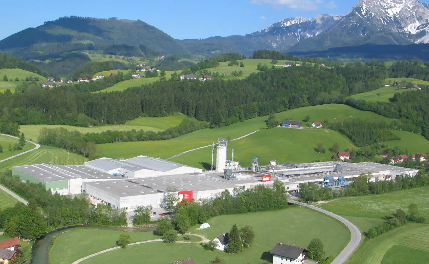 Luftaufnahme eines großen Industriegebäudes inmitten grüner Wiesen und Hügel mit Bergen im Hintergrund.