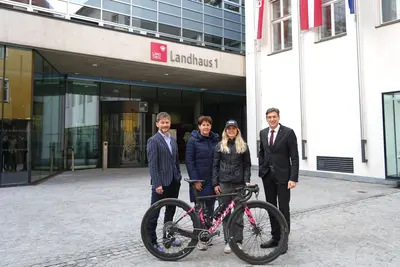 vor Gebäude vom Land Tirol, vier Personen stehen nebeneinander, von links nach rechts: Mann mit Sacko, Frau in Mantel, Frau mit Kappe und Jacke, Herr in Anzug und Krawatte, davor ein neues Fahrrad, Blick in Richtung Kamera