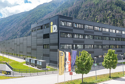 Ein großes modernes Gebäude mit mehreren Stockwerken vor einem bewaldeten Berghang. Vor dem Gebäude stehen mehrere Fahnenmasten mit Flaggen.