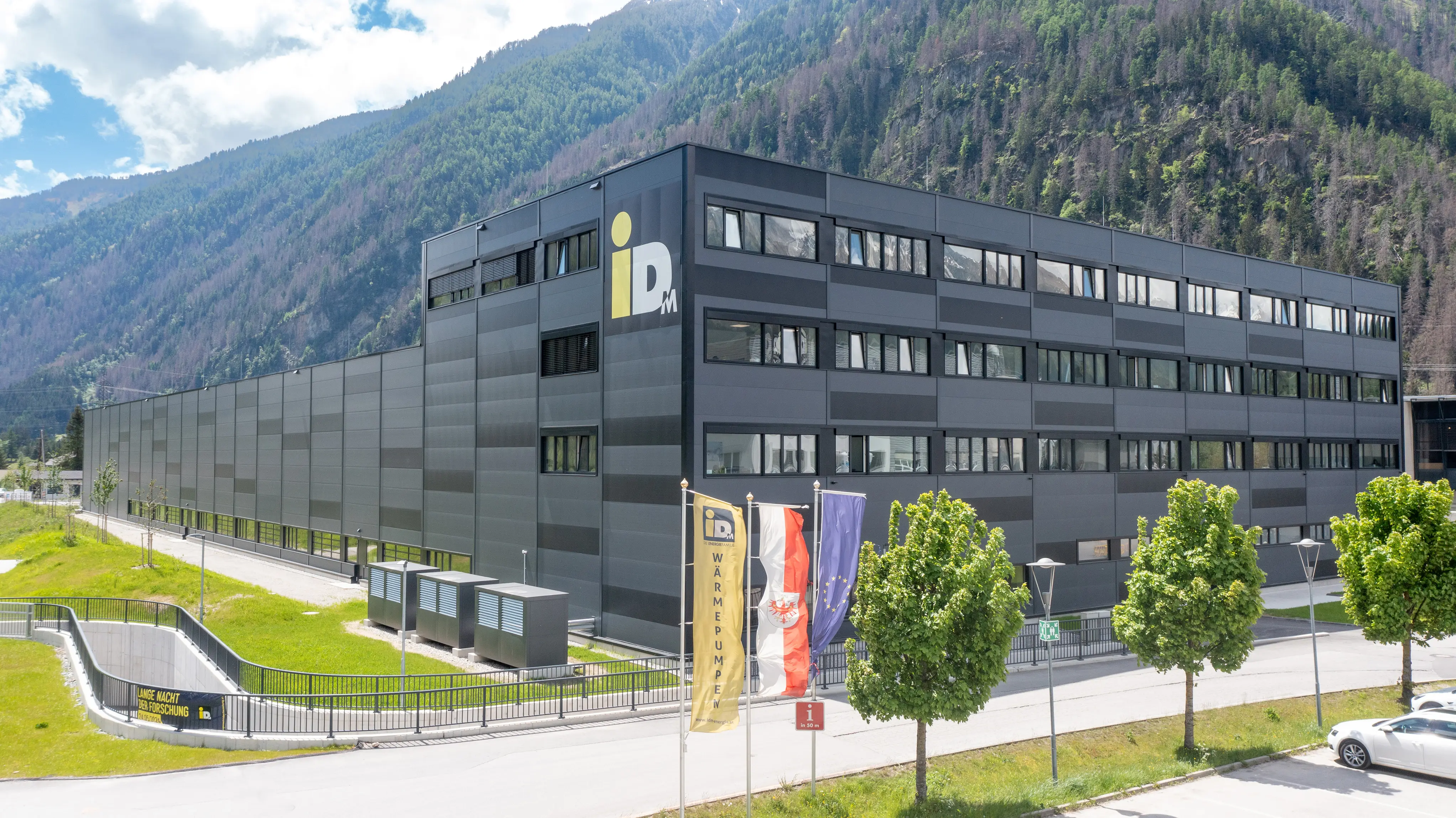 Ein großes modernes Gebäude mit mehreren Stockwerken vor einem bewaldeten Berghang. Vor dem Gebäude stehen mehrere Fahnenmasten mit Flaggen.