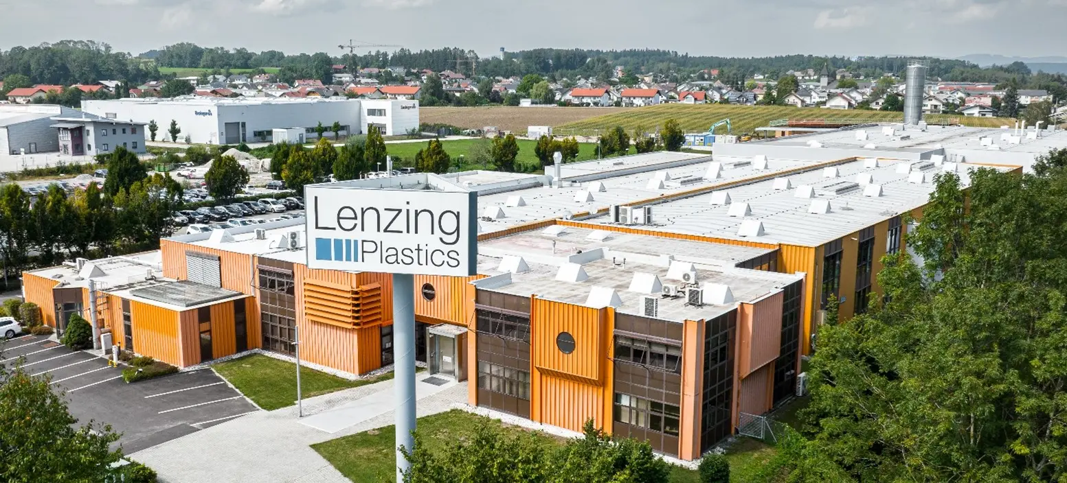 Luftaufnahme eines großen Industriekomplexes mit orangefarbenen Fassadenelementen und weißem Flachdach. Im Vordergrund ein Schild mit der Aufschrift „Lenzing Plastics“, im Hintergrund weitere Gebäude und Grünflächen