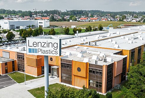 Luftaufnahme eines großen Industriekomplexes mit orangefarbenen Fassadenelementen und weißem Flachdach. Im Vordergrund ein Schild mit der Aufschrift „Lenzing Plastics“, im Hintergrund weitere Gebäude und Grünflächen
