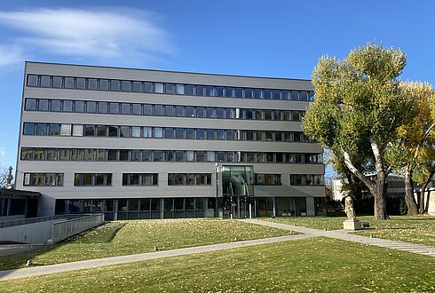 Ein modernes mehrstöckiges Bürogebäude mit großen Fenstern, umgeben von einer grünen Wiese und Bäumen unter klarem blauem Himmel.