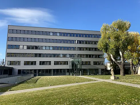 Ein modernes mehrstöckiges Bürogebäude mit großen Fenstern, umgeben von einer grünen Wiese und Bäumen unter klarem blauem Himmel.