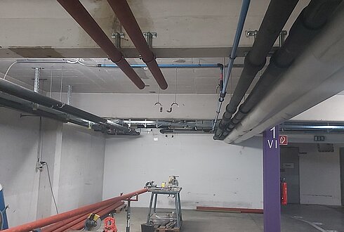 Mehrere farbige Rohrleitungen verlaufen entlang einer Betonwand in einem technischen Installationsbereich.