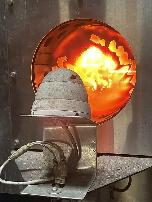 Nahaufnahme eines Industrieofens oder Brennofens mit sichtbarer Flamme im Inneren. Der Ofen besteht aus Metall und hat eine kreisförmige Öffnung, durch die das helle, intensive Feuer zu sehen ist. Vor der Öffnung ist ein Gerät auf einer Metallhalterung montiert, das über Kabel angeschlossen ist – vermutlich ein Sensor oder eine Kamera zur Überwachung der Ofenbedingungen.