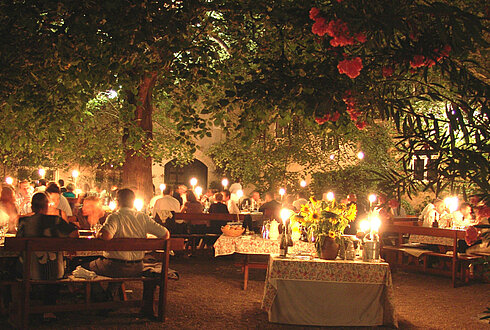 Abendstimmung an der Kaiserlinde im Nikolaihof Wachau. Zahlreiche Gäste sitzen bei stimmungsvoller Beleuchtung in entspannter Atmospähre unter einem dichten Blätterdach.