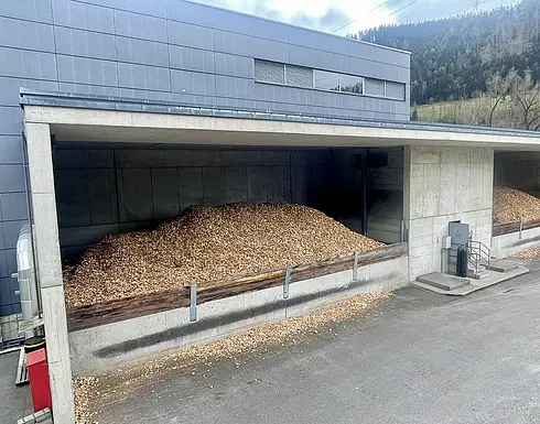 Lagerbereich mit zwei großen Holzschnitzelhaufen. Der Bereich gehört zu einem größeren Industriegebäude mit blauer Außenfassade und Betonwänden. Die Holzschnitzel befinden sich in getrennten Kammern mit Betonboden und -wänden, deren Vorderseiten teilweise offen sind und durch Metallbarrieren gesichert werden.