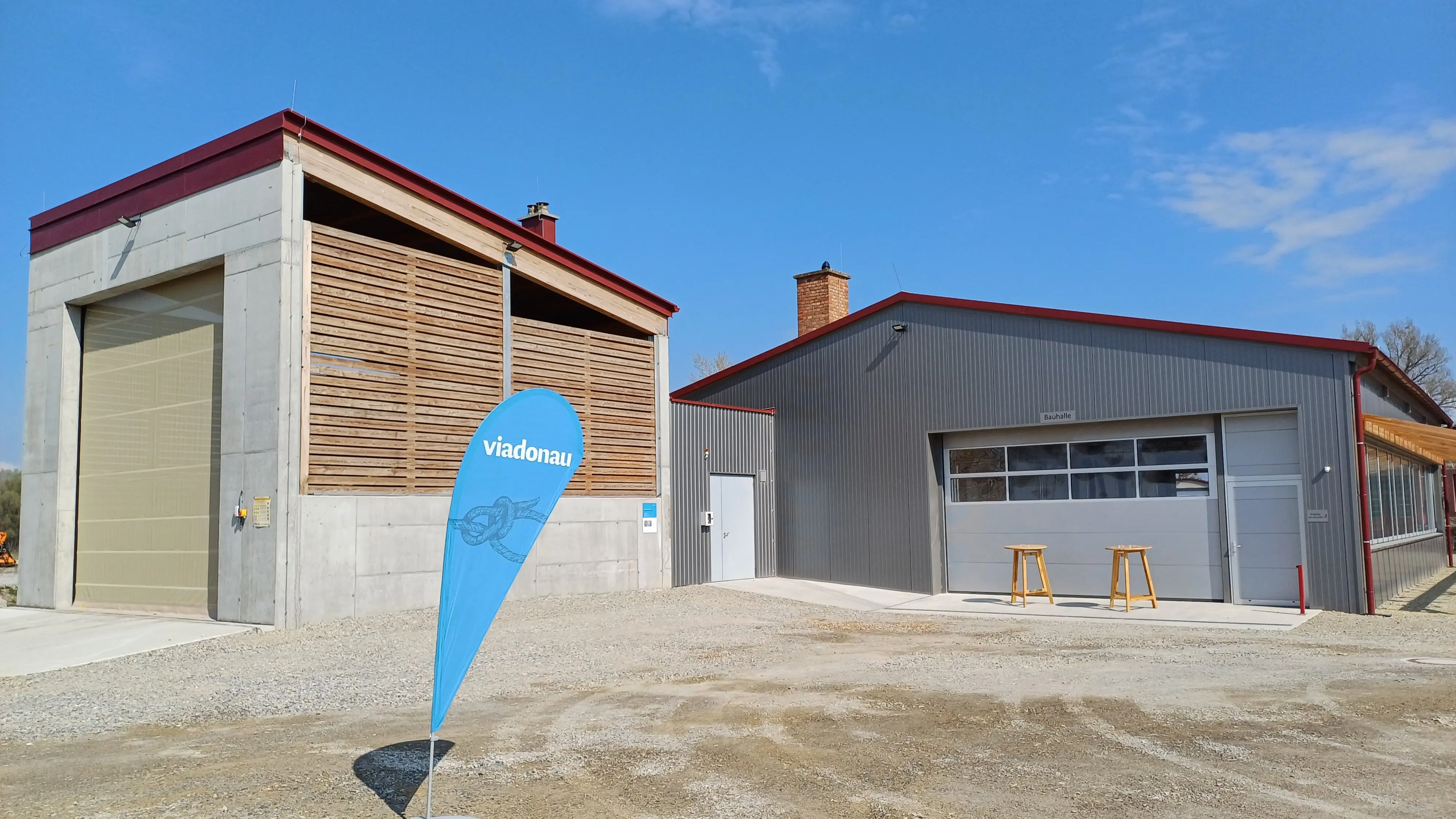 Zwei Gebäude mit Holzlamellen und Metallfassade auf einem Schotterplatz, davor ein blaues Banner mit der Aufschrift „viadonau“.