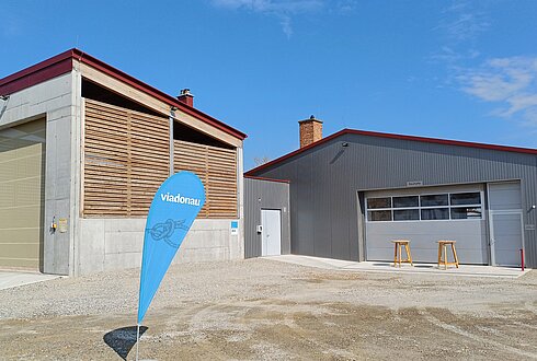 Zwei Gebäude mit Holzlamellen und Metallfassade auf einem Schotterplatz, davor ein blaues Banner mit der Aufschrift „viadonau“.