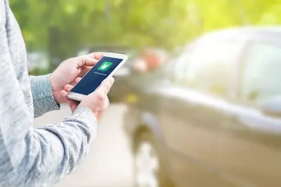 Hand hält Handy und eine App ist darauf zu sehen. Im Hintergrund ein Auto