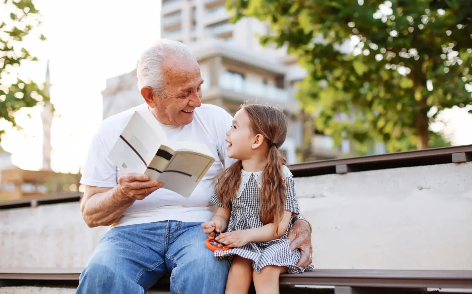 Großvater sitzt mit der Enkelin auf einer Sitzfläche im Freien. Er hält ein Buch.
