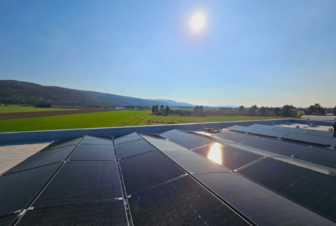 Solarmodule auf einem Flachdach, im Hintergrund grüne Felder und Hügel unter klarem blauem Himmel mit strahlender Sonne.