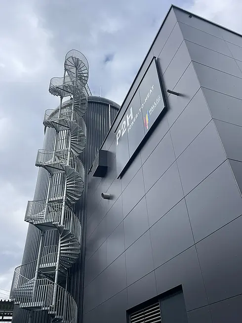 Modernes Industriegebäude mit einer großen zylindrischen Struktur, die über eine außenliegende Wendeltreppe aus Metall verfügt. Auf dem Gebäude befindet sich ein Schild mit der Aufschrift „P2H Power-to-Heat“ sowie dem Logo des Fraunhofer IEE. Im Hintergrund ist ein bewölkter Himmel zu sehen.