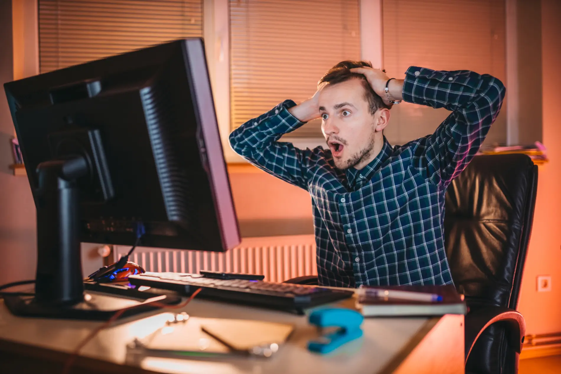 Ein Mann sitzt auf seinem Schreibtischsessel und hält erschrocken seine Hände an den Kopf. Er schaut dabei in seinen Monitorbildschirm