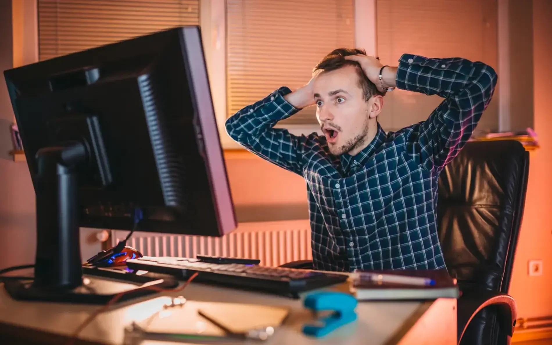 Ein Mann sitzt auf seinem Schreibtischsessel und hält erschrocken seine Hände an den Kopf. Er schaut dabei in seinen Monitorbildschirm