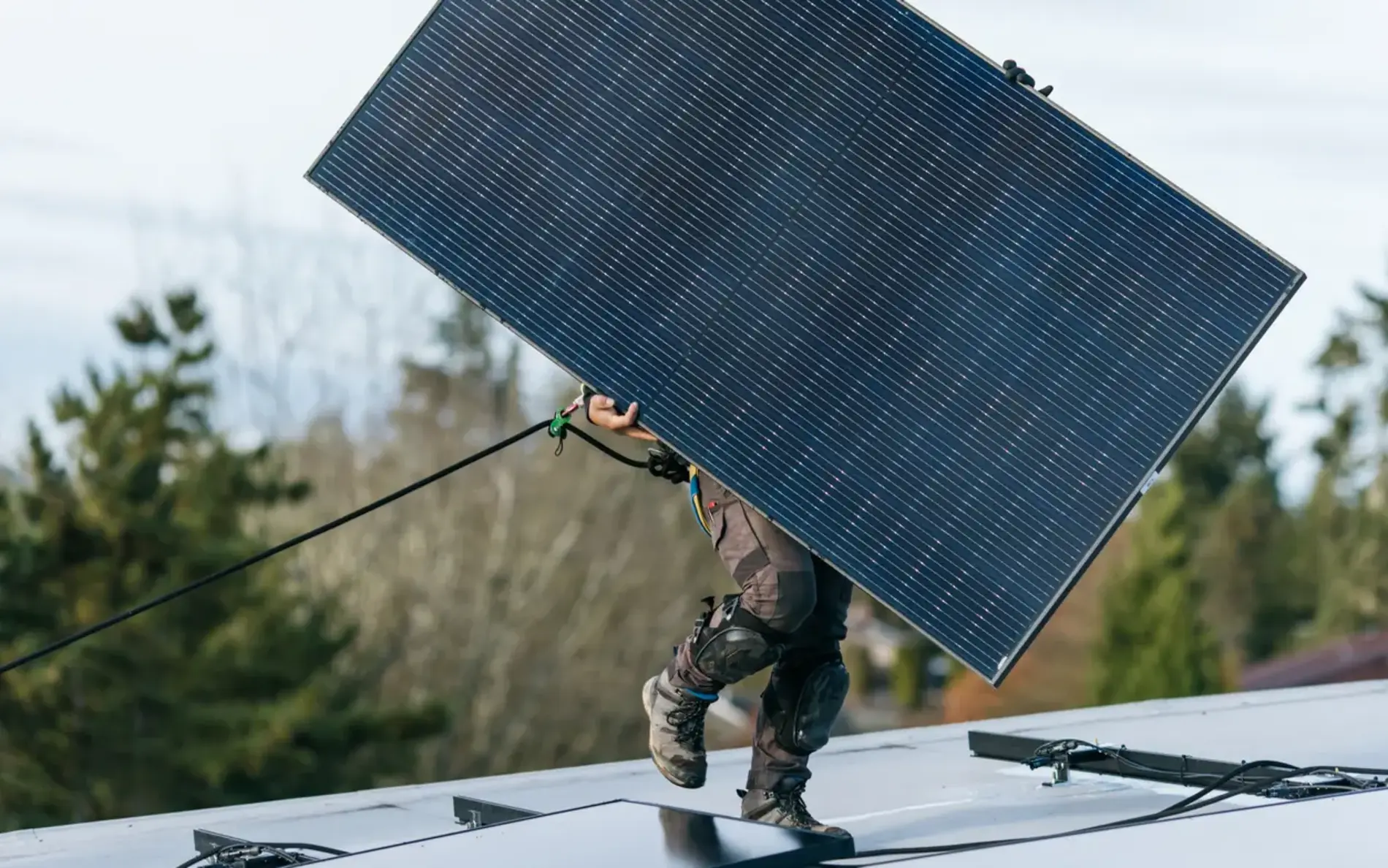 Ein PV Installateur trägt ein PV Paneel auf einem Dach. Er ist dabei mit einem Seil gesichert und trägt Knieschützer