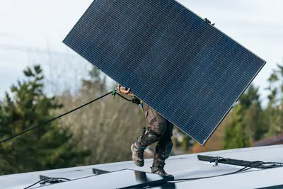 Ein PV Installateur trägt ein PV Paneel auf einem Dach. Er ist dabei mit einem Seil gesichert und trägt Knieschützer