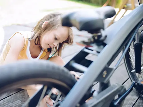 Mädchen sitzt am Boden vor ihrem Fahrrad. Man sieht durch die Speichen, wie sei ein Werkzeug hält und versucht es zu reparieren.