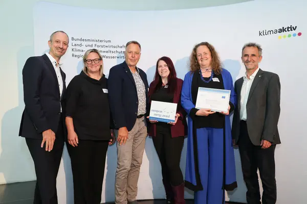 Menschen mit einer Urkunde in der Hand vor einer Fotowand. Sie lächeln in die Kamera.