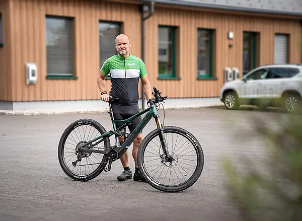 Mann steht hinter Fahrrad, trägt schwarz-grünes Fahrradtrikot mit Aufschrift ÖkoFEN, im Hintergrund Gebäude mit elektrischem Auto