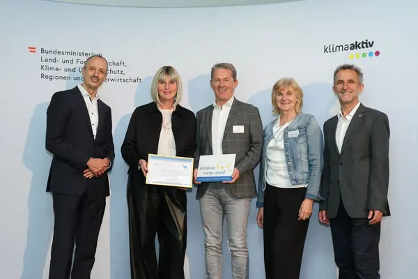 Menschen mit einer Urkunde in der Hand vor einer Fotowand. Sie lächeln in die Kamera.