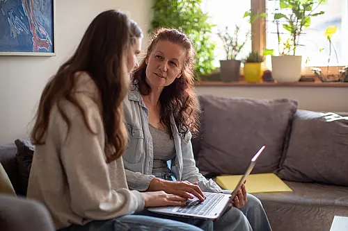 Zwei Frauen sitzen mit einem Laptop auf der Couch, die eine Erklärt der anderen was.