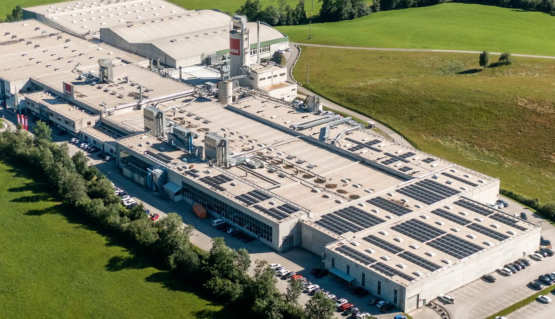 Luftaufnahme einer großen Produktionshalle mit installierten Solarmodulen auf dem Dach und angrenzenden Parkflächen.