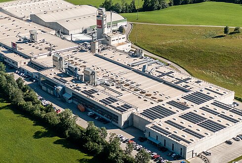 Luftaufnahme einer großen Produktionshalle mit installierten Solarmodulen auf dem Dach und angrenzenden Parkflächen.