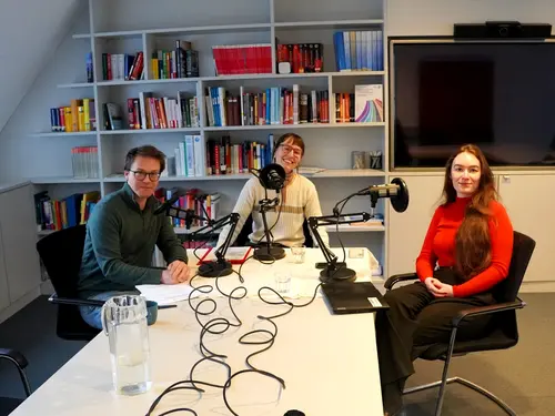 3 Menschen lächeln in die Kamera. Vor ihnen stehen Podcast-Mikrofone.