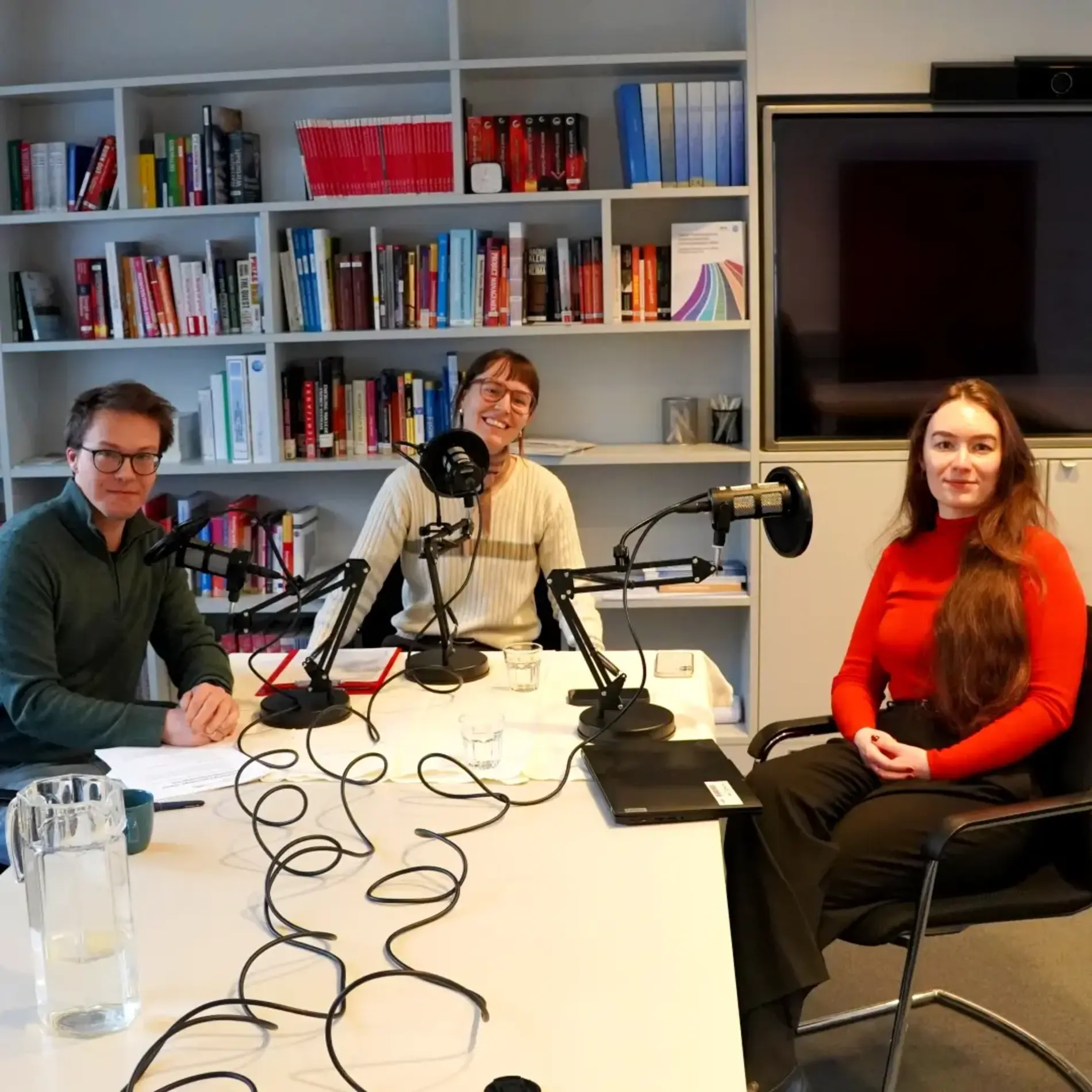 3 Menschen lächeln in die Kamera. Vor ihnen stehen Podcast-Mikrofone.