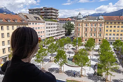 Blick von einem erhöhten Standort auf den Bozner Platz in Innsbruck. Im Vordergrund blickt eine Person über den Platz. Der neu gestaltete Platz ist mit zahlreichen jungen Bäumen und Sitzgelegenheiten begrünt; in der Mitte steht ein Brunnen. Mehrgeschossige Gebäude umgeben den Platz, im Hintergrund sind die Berge rund um Innsbruck zu sehen.