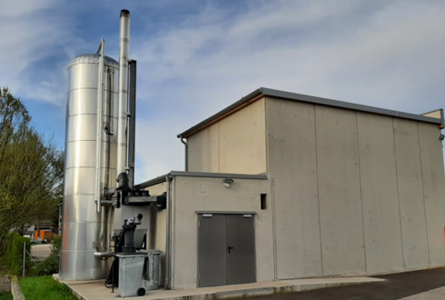 Industriegebäude mit grauen Betonwänden und einem hohen, silberfarbenen Zylindertank mit Rohrleitungen und Schornstein, vermutlich Teil einer Biomasse- oder Heizungsanlage