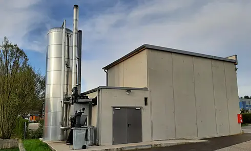 Industriegebäude mit grauen Betonwänden und einem hohen, silberfarbenen Zylindertank mit Rohrleitungen und Schornstein, vermutlich Teil einer Biomasse- oder Heizungsanlage