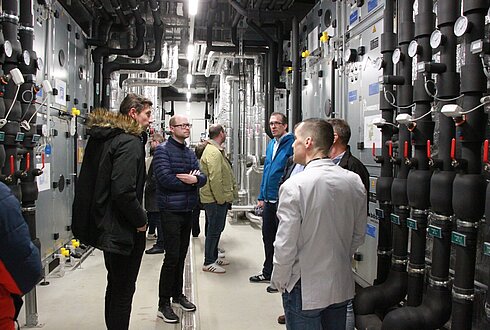 Exkursions-Teilnehmer:innen bei der Besichtigung der Energiezentrale, Landeskrankenhaus Hall. Mehrere Personen stehen in einem Raum mit technischen Anlagen, darunter Rohrleitungen, Messgeräte und Steuerungselemente. Die Personen tragen Alltags- und Businesskleidung, ihre Gesichter sind unkenntlich gemacht.