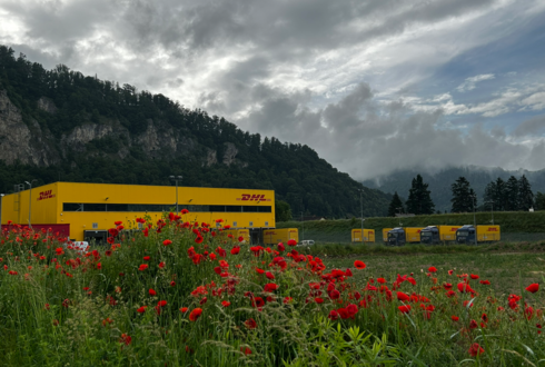 DHL-Logistikzentrum in gelber Farbe vor bewaldeten Hügeln, mit roten Mohnblumen im Vordergrund und mehreren DHL-Lkw im Hintergrund unter bewölktem Himmel.