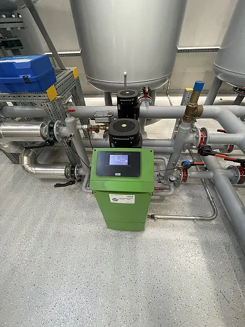 Technikanlage mit grünem Steuergerät und mehreren Leitungen, Pumpen und Ventilen in einem Gebäudetechnikraum.