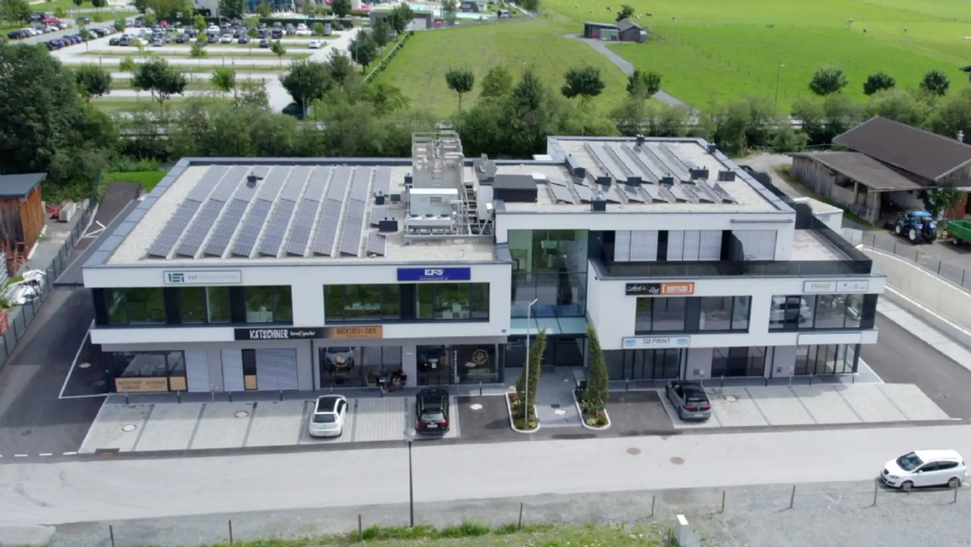 Luftaufnahme vom G7-Gewerbepark in Kaprun. Ein flaches Gebäude mit einer PV-Installation auf dem Dach, einem schmalen Parkplatz und einer Wiesenlandschaft im Hintergrund.