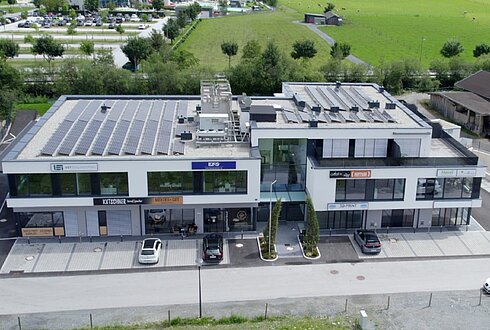 Luftaufnahme vom G7-Gewerbepark in Kaprun. Ein flaches Gebäude mit einer PV-Installation auf dem Dach, einem schmalen Parkplatz und einer Wiesenlandschaft im Hintergrund.
