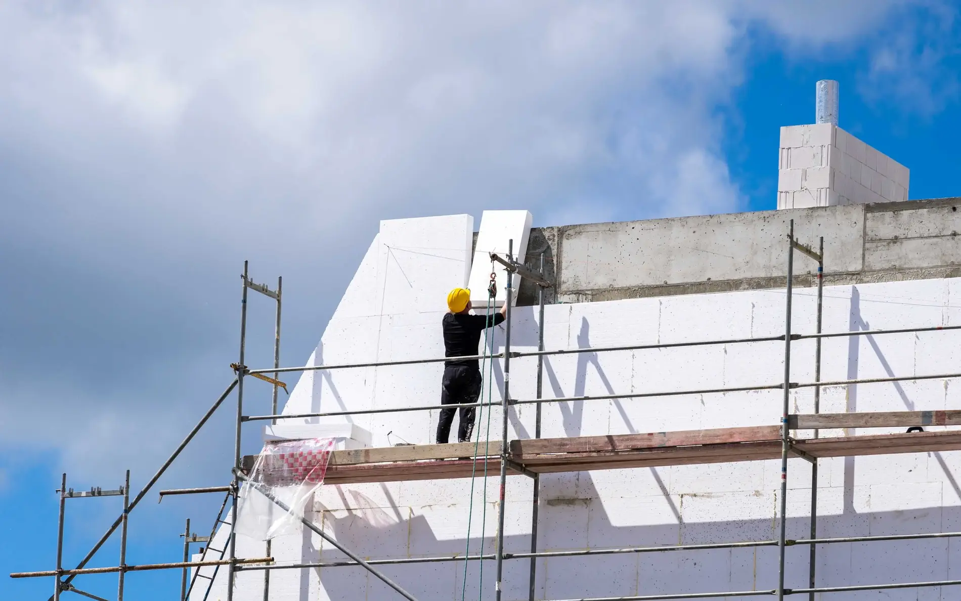 Ein Gerüst um ein Haus, das gerade saniert wird. Auf den Wänden sind Dämmplatten installiert. Ein Mann steht oben am Gerüst und installiert die letzten Dämmplatten.