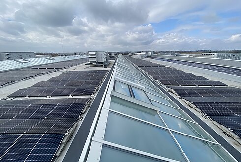 Solarmodule auf einem Dach neben langen Glas-Lichtbändern, die Tageslicht in die Produktionshalle bringen.