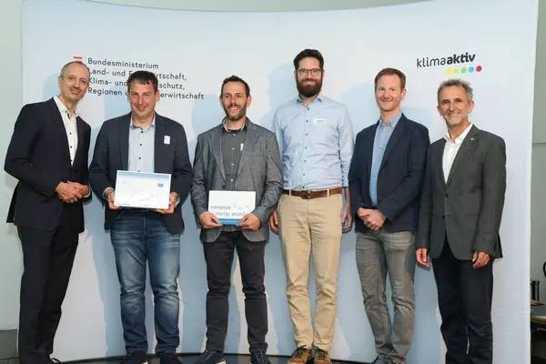 Menschen mit einer Urkunde in der Hand vor einer Fotowand. Sie lächeln in die Kamera.