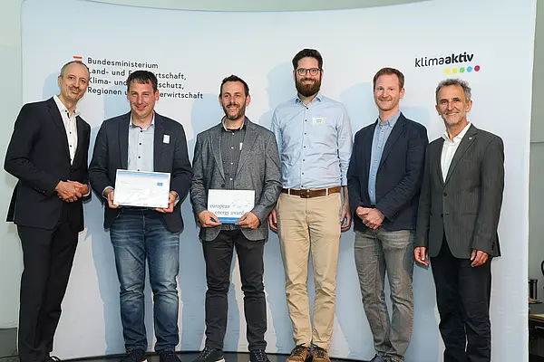 Menschen mit einer Urkunde in der Hand vor einer Fotowand. Sie lächeln in die Kamera.