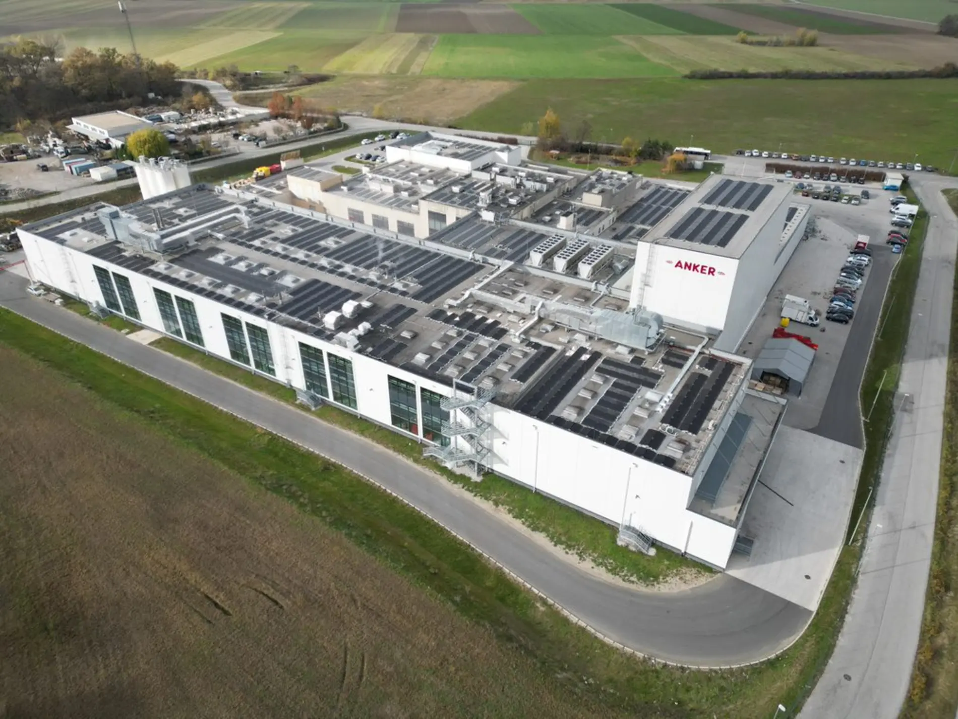 Luftaufnahme eines großen Industriegebäudes mit mehreren Dachinstallationen, darunter vermutlich Solarpaneele und Lüftungssysteme. Das Gebäude liegt inmitten von Feldern, ist von einer Straße umgeben und trägt die Aufschrift „ANKER“. Mehrere Fahrzeuge sind auf dem Gelände geparkt.
