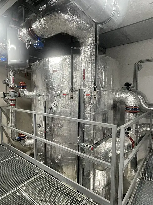 Industrielle Anlage mit isolierten Rohrleitungen, Tanks, Ventilen und Messgeräten – vermutlich Teil eines Heizungs- oder Lüftungssystems. Metallgeländer und Gitterboden sind sichtbar.